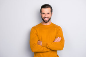 Portrait of attractive self-assured with beaming shiny smile haircut with long furfur wearing casual classic color of mustard sweater macho man standing with folded arms isolated on gray background