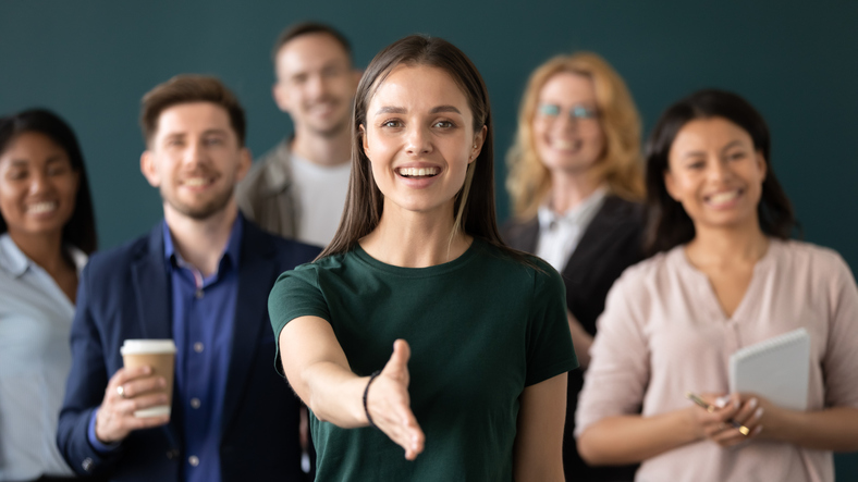 Friendly young company woman representative holds out her hand for handshake welcoming customer smiling looking at camera posing together with diverse colleagues