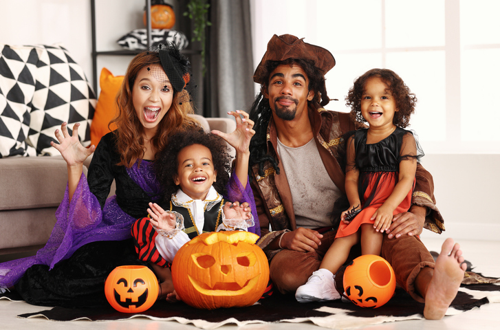Joyful family parents and children in Halloween costumes making scary gesture and smiling at camera while sitting on with jack-o-lantern on floor, celebrating all hallows day