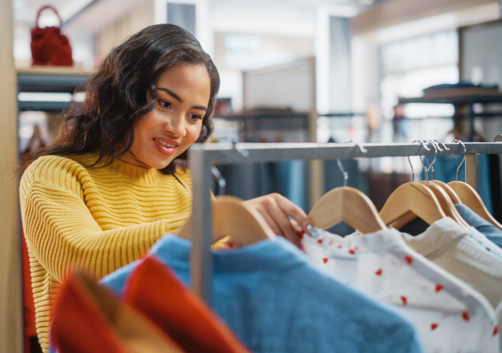 Young woman in yellow sweater shopping clothing racks in retail store.